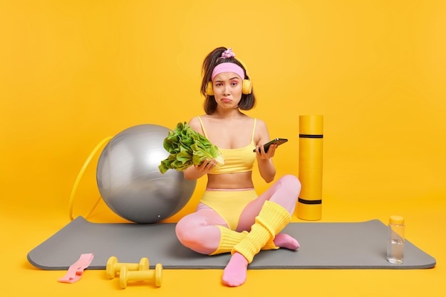 Displeased Asian woman dressed in sportswear leads healthy lifestyle holds lettuce salad and smartphone poses on fitness mat feels hesitant uses dumbbells resistance band hula hopp and fitball