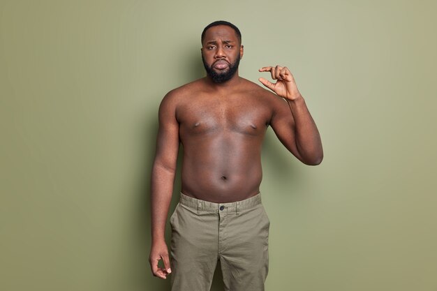 Displeased bearded Afro American man poses with naked torso shows tiny little thing gesture displays very small object poses against dark green wall