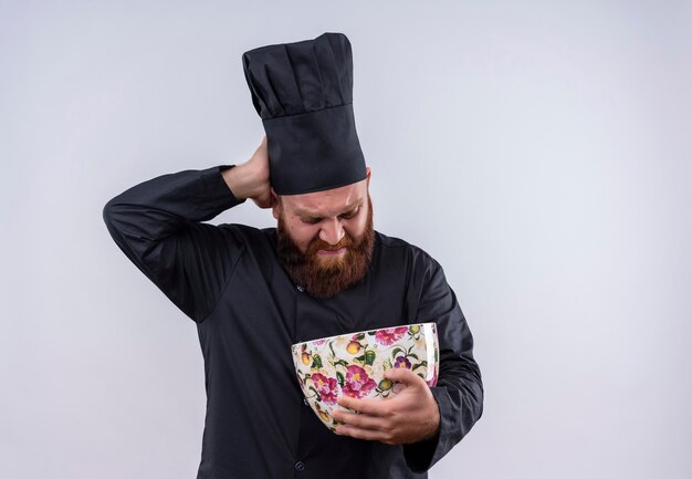 A displeased bearded chef man in black uniform holding huge floral cup and looking at it with negative expression on a white wall