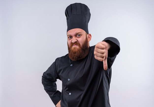 Недовольный бородатый шеф-повар в черной униформе показывает палец вниз и смотрит на белую стену