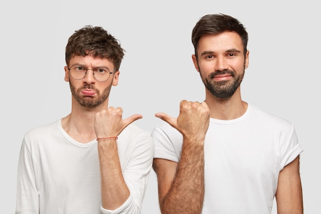 Displeased bearded male feels offended of bad jokes of his friend, point at each other, argue about something, dressed in casual white t-shirts. People, relationship, friendship