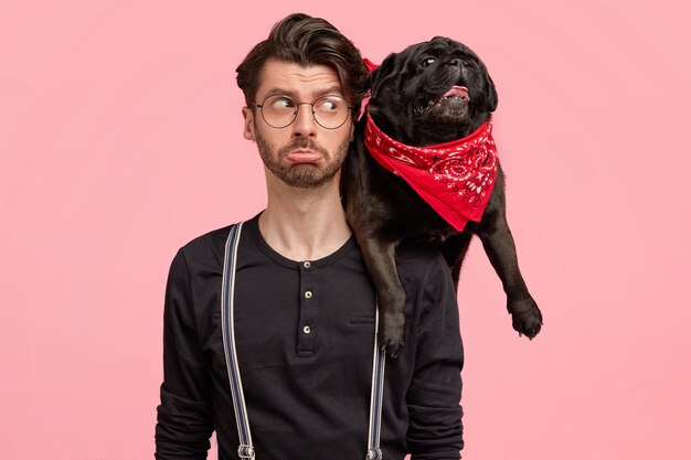 Displeased bearded male purses lips, looks with negative facial expression at his dog, being discontent after walk, pose together against pink wall. People, animals, relationships, lifestyle