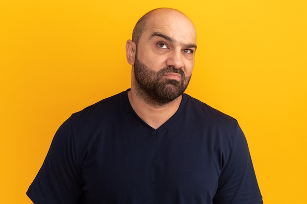 Uomo barbuto scontento in maglietta blu scuro che osserva in su facendo la bocca ironica con espressione delusa in piedi sopra la parete arancione