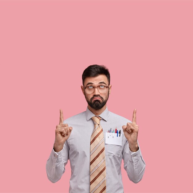 Free photo displeased bearded young man frowns face, points with both index finger upwards, being doubtful, feels uncertain