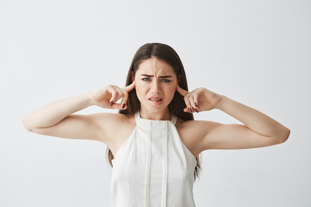 Displeased beautiful brunette girl closing ears with fingers from noise biting lip .