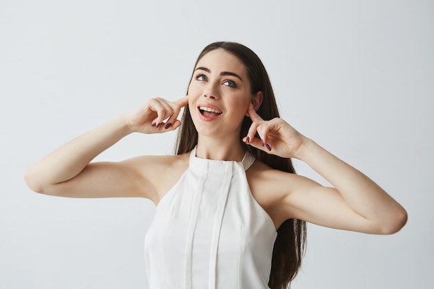 Displeased beautiful brunette girl closing ears with fingers from noise smiling with opened mouth .