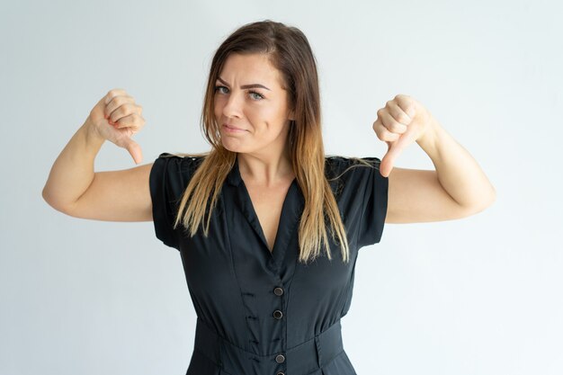 Displeased beautiful woman in black dress showing thumbs down and looking at camera. 