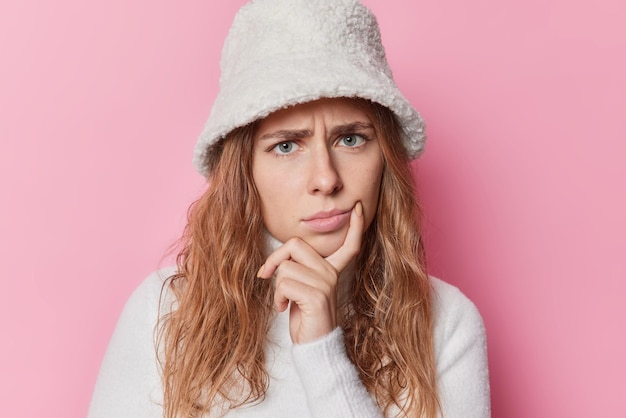 La bella donna dispiaciuta tiene il dito vicino all'angolo delle labbra sembra sconvolto concentrato con uno sguardo triste attento indossa panama invernale alla moda e maglione isolato su sfondo rosa. emozioni negative