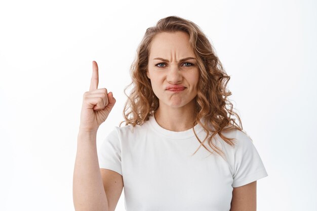 Displeased blond woman grimacing looking disappointed and skeptical pointing finger up at something bad and upsetting complaining white background
