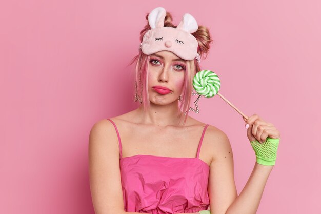 Displeased blonde young European woman with bright makeup holds lollipop looks sadly at camera holds green sweet