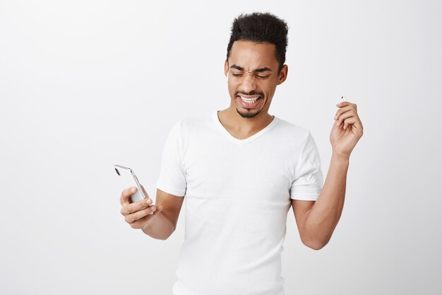 Недовольный и обеспокоенный афро-американский парень снимает наушники с громкого ужасного шума, останавливает музыку на мобильном телефоне