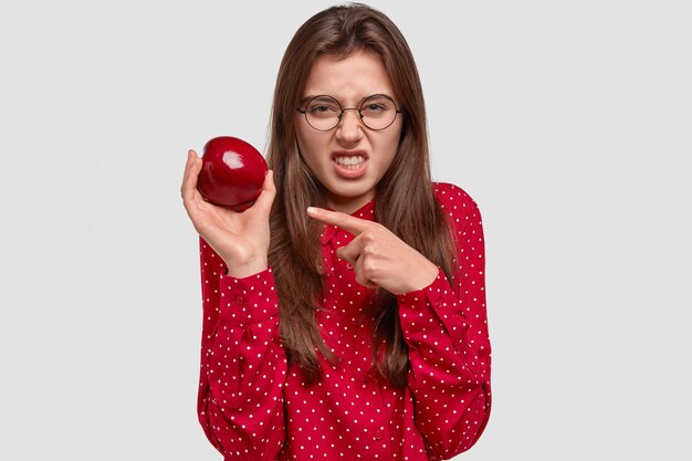 Displeased brunette woman clenches teeth, frowns face in discontent, holds apple, doesnt like its taste