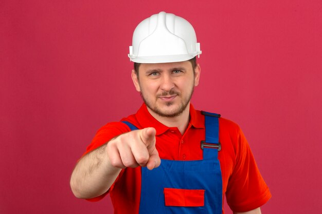 Displeased builder man wearing construction uniform and security helmet pointing with finger to camera over isolated pink wall