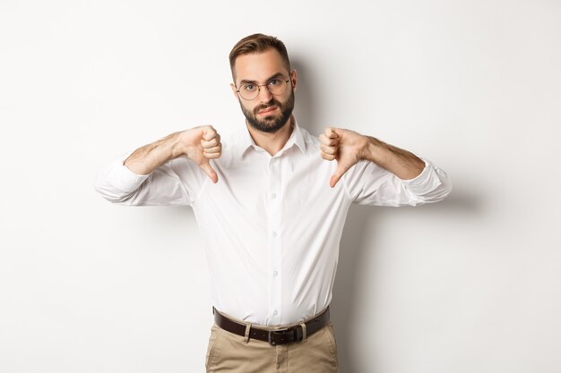 Displeased businessman in glasses showing thumbs down, dislike and disapprove, standing  white