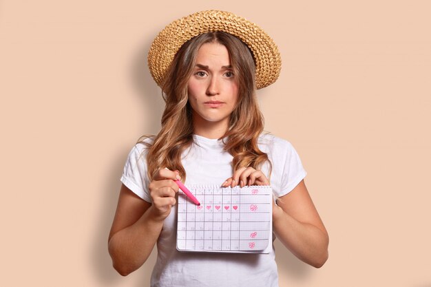 Недовольная кавказская женщина в повседневной футболке и соломенной шляпе, указывает на календарь периода, не хочет менструации во время отдыха на берегу моря, изолированная на бежевом. Несчастная крытая женщина