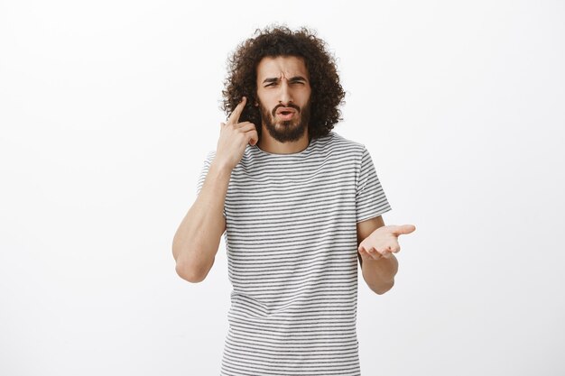 displeased confused bearded male student in striped t-shirt, pointing at ear and frowning, cannot hear question while being in loud company