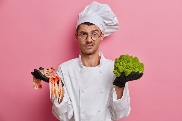 Displeased cook in white uniform, works at restaurant, given task to cook dish from broccoli and crayfish, wears black gloves