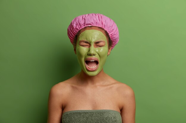 Displeased crying woman feels tired of beauty treatments in spa salon, keeps mouth opened, wears green mask for perfect skin, wears bath cap and towel around body, stands against  wall.