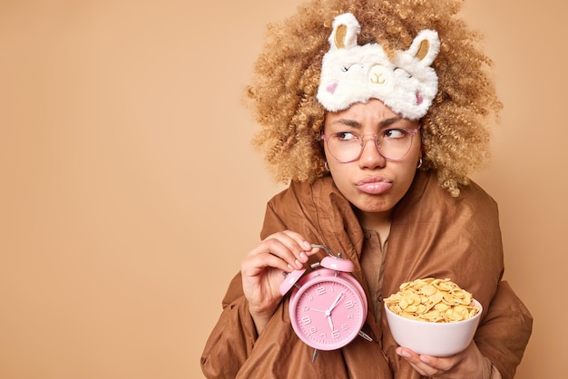 Free photo displeased curly haired woman wrapped in blanket holds bowl of cornflakes and alarmclock has gloomy expression wears sleepmask on forehead isolated over brown background copy space for your promo