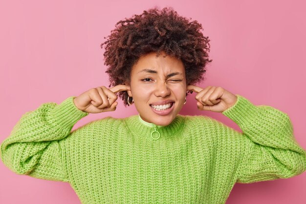 Displeased curly haired young woman plugs ears cannot focus in crowded place feels discomfort over noise being troubled to listen advice wears green knitted sweater isolated over pink studio wall