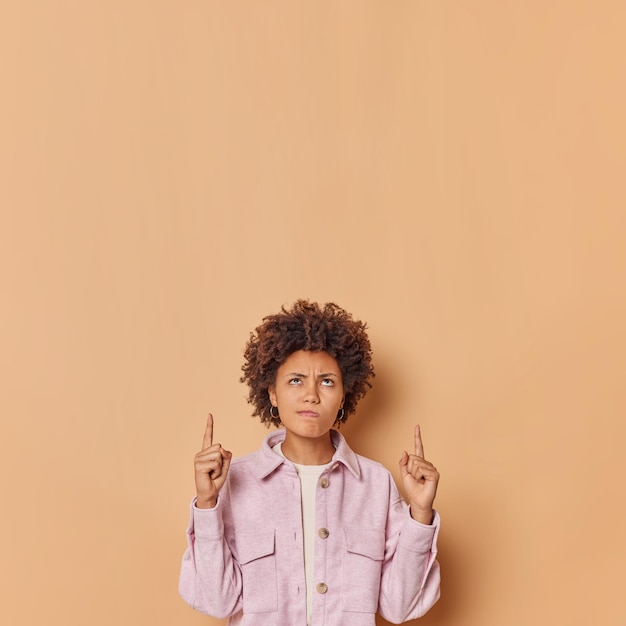 Displeased curly haired young woman points above with both index fingers dissatisfied with suggested sale discount purses lips looks angrily unhappily wears jacket isolated over brown background