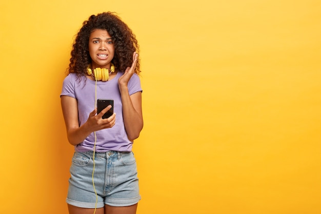Displeased dark skinned curly woman listens disgusting voice message in headphones, holds modern smartphone, wears purple t shirt and denim shorts, stands against yellow studio wall, copy space