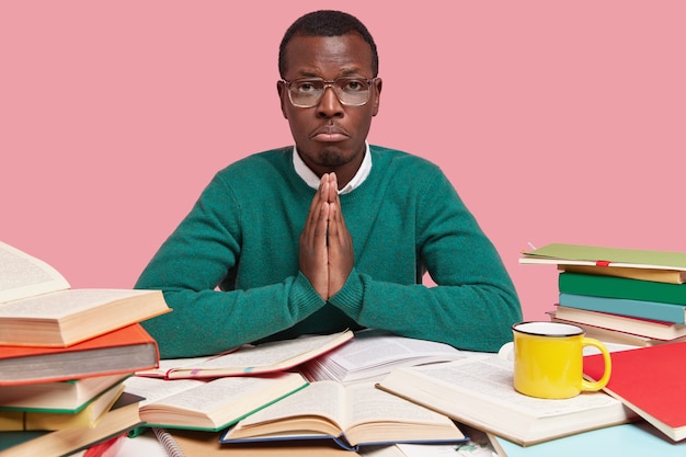 Displeased dark skinned man has sorrowful expression, keeps palms in praying gesture, believes in good luck while passing exam