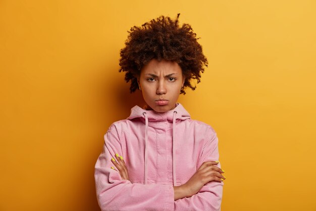 Displeased dark skinned woman stands in closed posture, smirks and sulks from anger