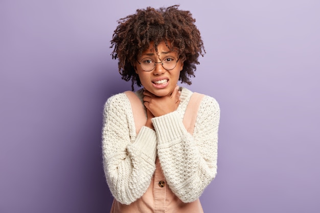 Displeased dark skinned woman suffers from suffocation, keeps both hands on throat, clenches teeth, has curly Afro hair