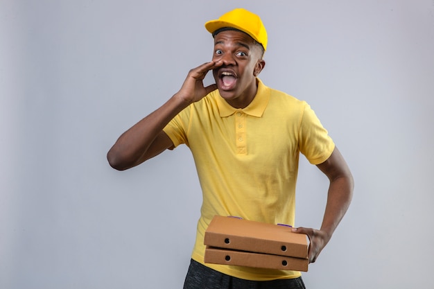 Displeased delivery african american man in yellow polo shirt and cap holding pizza boxes shouting with hand near mouth standing