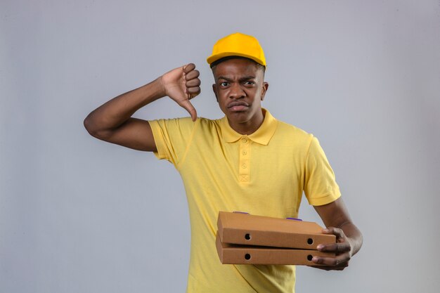 Displeased delivery african american man in yellow polo shirt and cap holding pizza boxes showing thumb down standing