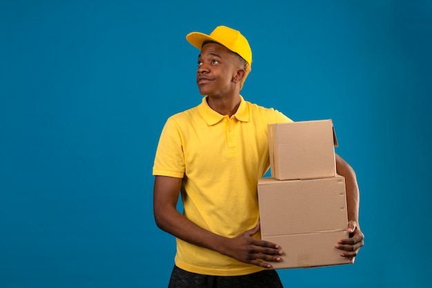 Displeased delivery african american man in yellow polo shirt and cap standing with cardboard boxes looking aside on isolated blue