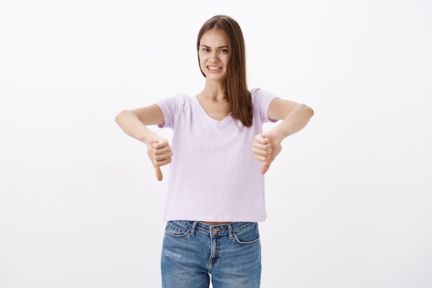 недовольная недовольная привлекательная молодая женщина-покупательница в блузке и джинсах показывает палец вниз, неловко улыбаясь, давая отрицательный отзыв и отвергая плохую идею над белой стеной