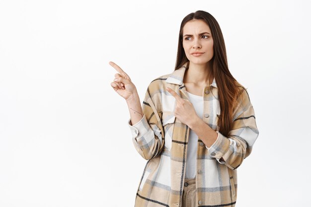 Displeased and doubtful female client, looking aside and frowning skeptical, having doubts with product quality, standing disappointed against white wall