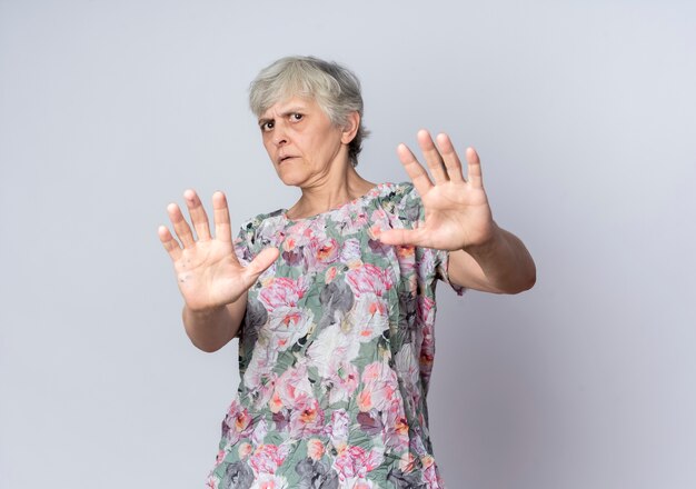 Displeased elderly woman defends holding out hands isolated on white wall