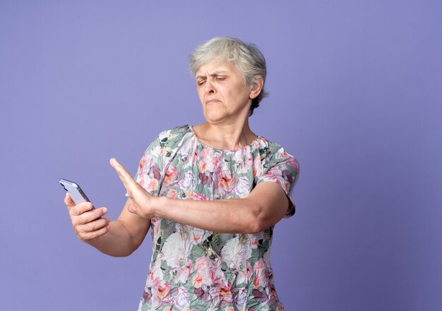 Displeased elderly woman holds and pretends to push phone isolated on purple wall