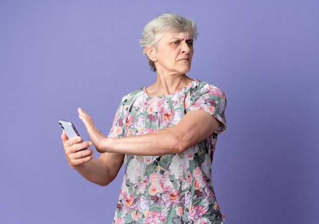 Displeased elderly woman holds and pretends to push phone looking at side isolated on purple wall