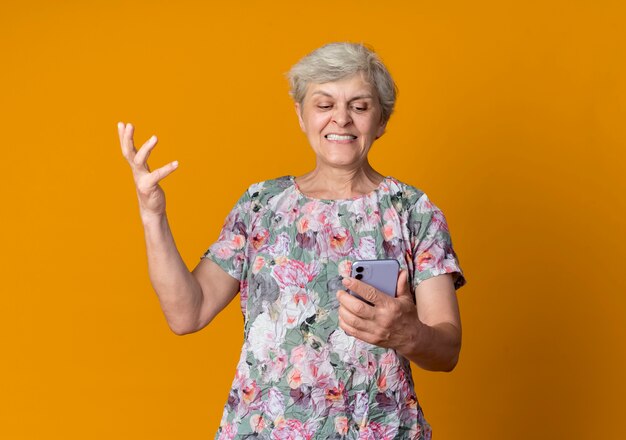 Displeased elderly woman raises hand holding and looking at phone isolated on orange wall