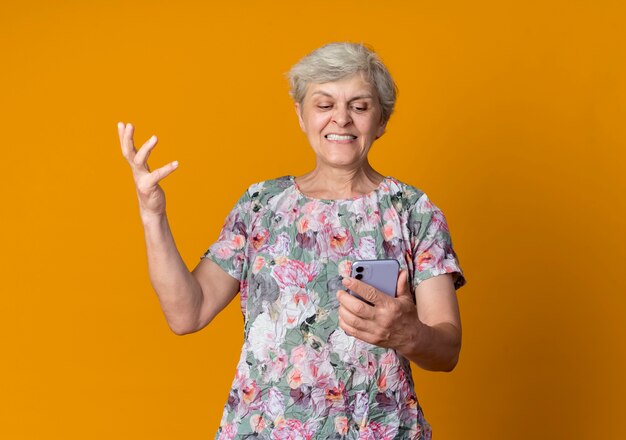 Недовольная пожилая женщина поднимает руку, держа и глядя на телефон, изолированный на оранжевой стене