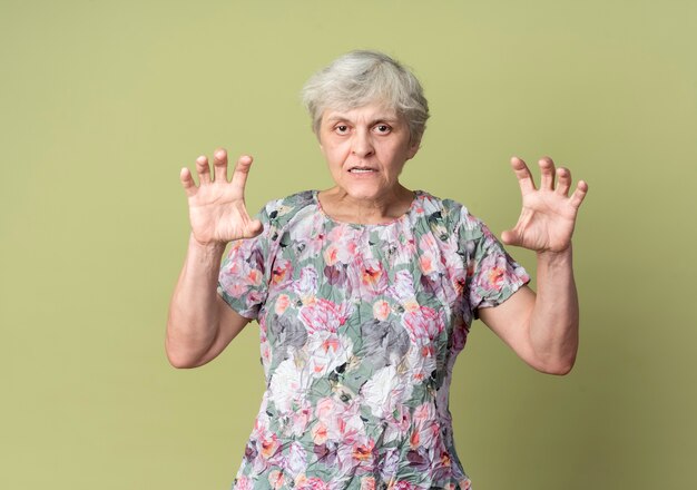 不機嫌な年配の女性は、オリーブグリーンの壁に隔離された手を上げて握り締めます