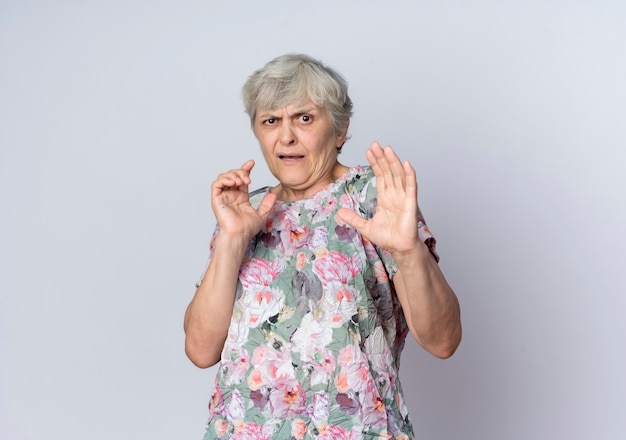 Displeased elderly woman stands with raised hands isolated on white wall