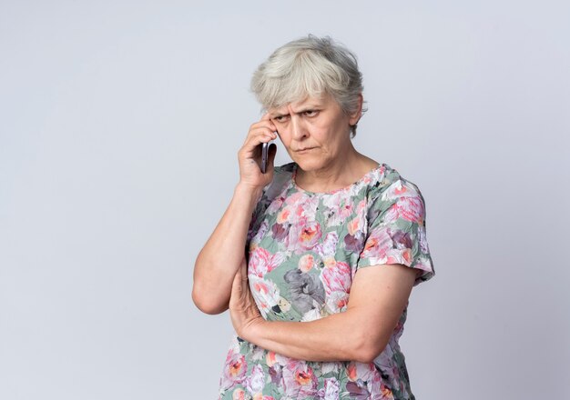 Displeased elderly woman talks on phone isolated on white wall