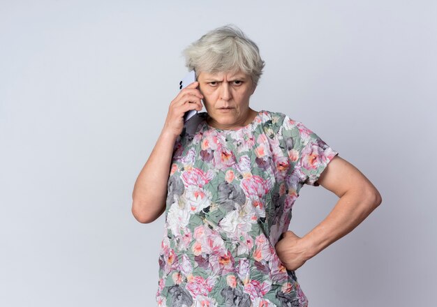 Раздраженная пожилая женщина разговаривает по телефону, изолированному на белой стене