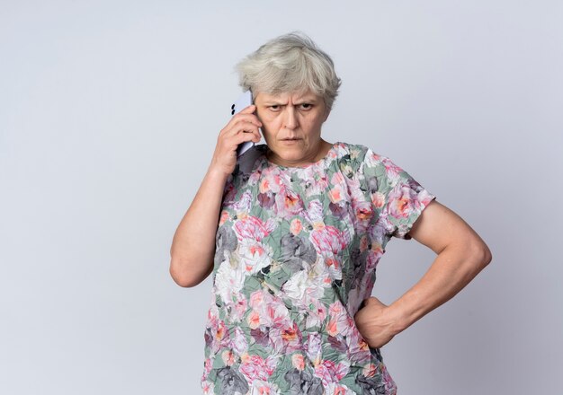 Displeased elderly woman talks on phone isolated on white wall