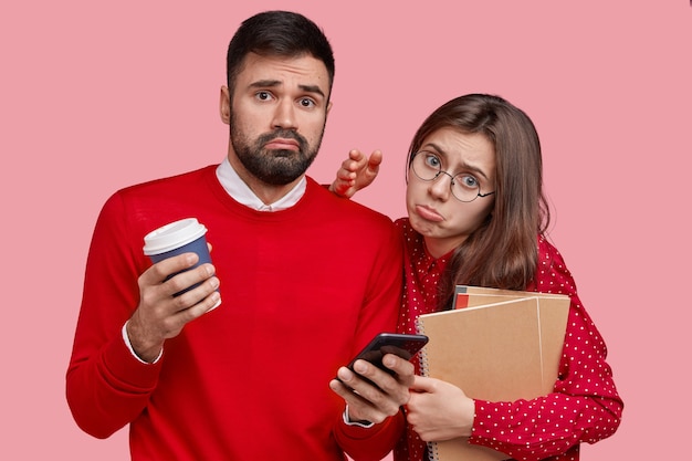 Displeased European lady and her boyfriend pose together, wear clothes in one colour have coffee break, use modern smart phone for online communication