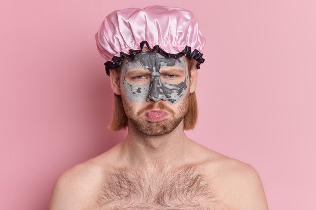 Displeased European man frowns face and looks with gloomy expression applies clay mask for skin refreshing wears bath hat stands topless