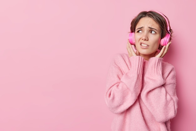 Displeased European with short dark hair keeps hands on stereo headphones doesnt like quality of sound looks unhappily away wears soft jumper isolated over pink background with copy space for text