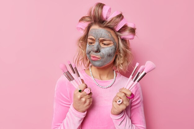 Displeased European woman applies clay mask on face chooses cosmetic brush going to do makeup wears hair rollers for making hairstyle dressed in festive outfit isolated over pink background