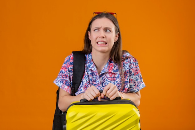 Displeased and fearful young traveler woman wearing red sunglasses on head with backpack holding suitcase over orange wall