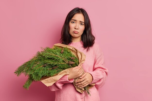 不機嫌な女性の花屋は、クリスマスリースをカメラで不幸に見えるようにするためにモミの木の枝を運びます家を飾るための美しい休日の構成を準備します長袖のジャンパーを着用します